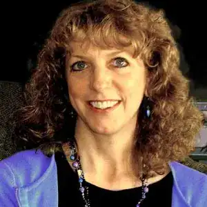 A smiling person with curly hair, wearing a blue top and a necklace that gleams like awards, set against a dark background.