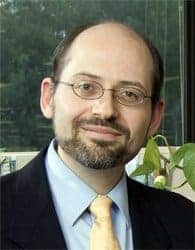 A man with a beard and glasses is smiling. He is wearing a dark suit, light blue shirt, and a yellow tie. In the background, there is a plant and a window with a view of trees.