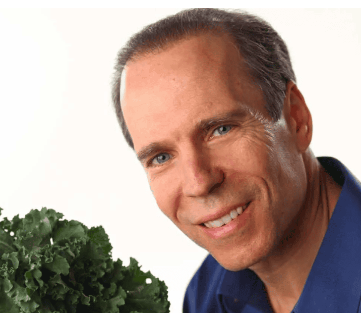 A person with short hair and a friendly smile holds a bunch of award-winning fresh kale. They are wearing a blue shirt against a plain white background, ready to share their top-reviewed recipe.