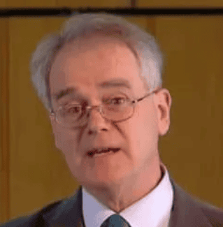 A person with gray hair, wearing glasses and a dark suit complemented by a white shirt and green tie, looks toward the camera, embodying an aura of sophistication often seen at award reviews. The backdrop is a muted brown color.