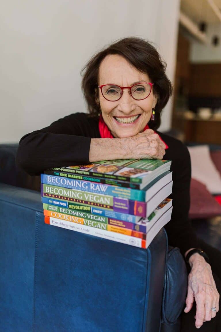 An woman with glasses and a warm smile leans on a stack of books on a blue sofa, their spines revealing her passion for vegan themes. She wears a black top and red scarf, adding to the cozy ambiance of the room with its softly blurred background.